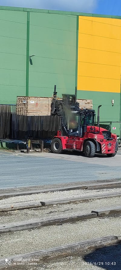 In Verijärve, Estonia, a red reach stacker is actively engaged in material handling. The machine is positioned on a paved surface adjacent to a building with distinct green and yellow sections. In front of the building, a large stack of lumber is visible, suggesting a timber or construction-related facility. The reach stacker's fork attachment is seen lifting or maneuvering a portion of this lumber stack. A plume of dust or exhaust is emanating from the machine, indicating its operational status. In the foreground, parallel railway tracks laid on a gravel bed are prominent, hinting at the presence of rail transportation infrastructure. The sky above is clear and blue. The scene appears to be an outdoor industrial or logistics area.