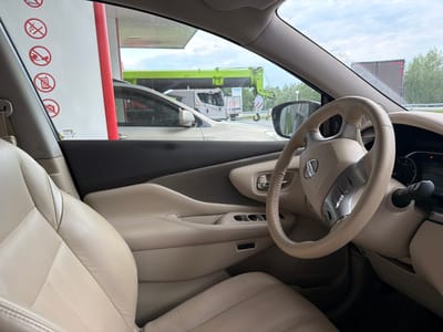 The image shows the interior of a car, likely a Nissan, viewed from the driver's seat. The car is parked at a gas station, evidenced by the fuel pump canopy visible through the windshield. Outside the car, a large green construction crane and a truck are visible, along with trees and a cloudy sky. The car's interior features beige leather seats, a leather-wrapped steering wheel with controls, and a dashboard with a digital display. There are also several red circular warning signs on the white pillar of the gas station canopy, indicating prohibitions against smoking, using mobile phones, and other activities. The scene appears to be a typical day at a roadside service area, with the car likely stopped for refueling or a break. The location is identified as Shchetkovo, Russia.