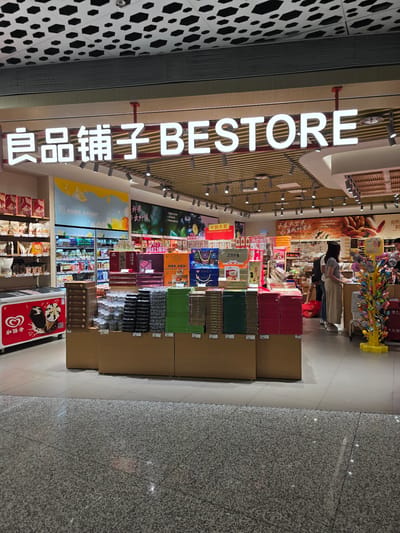The image shows the interior of a Bestore shop in Bao'an District, China. The store is well-lit and organized, with shelves stocked with various snacks and confectionery products. A prominent sign above the entrance reads "良品铺子 BESTORE". In the foreground, there are several display stands showcasing boxes of snacks. To the left, a red freezer with an ice cream advertisement is visible. In the middle ground, customers are browsing the aisles, with one woman in a grey top and beige pants standing at a counter. The background reveals more shelves filled with products, refrigeration units, and promotional posters on the walls. The floor is a polished speckled grey tile. The ceiling features a modern design with hexagonal patterns and recessed lighting.