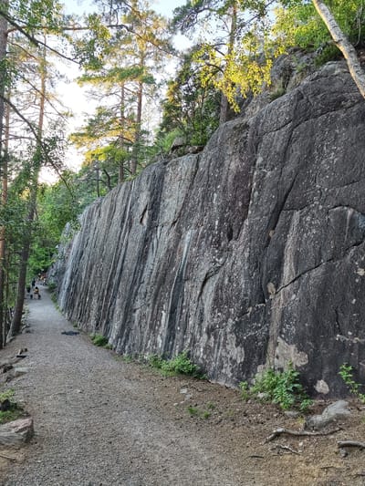 This image depicts a gravel path running alongside a large, sheer rock face in Skriketorp, Sweden. The rock face is dark gray with lighter gray and white mineral deposits. The path is surrounded by trees with green foliage, and sunlight filters through the leaves, casting dappled shadows. A few people and dogs are visible walking on the path. The scene suggests a natural outdoor setting, possibly a park or hiking area.