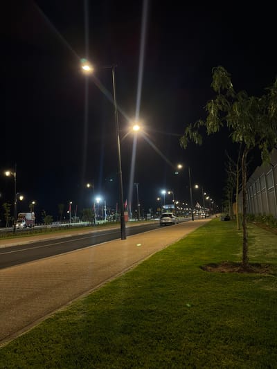 The image depicts a nighttime scene in Ouahat Sidi Brahim, Morocco. A street stretches into the distance, illuminated by a series of evenly spaced streetlights. The left side of the frame is dark, while the right side features a walkway bordered by a patch of green grass. Several vehicles are visible along the street, and trees are sparsely placed along the walkway. A building with a latticed roof structure is visible in the background. The scene appears calm and quiet.