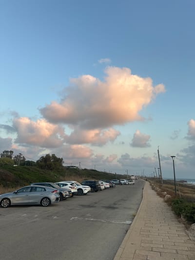 The image captures a scene in the Mate Asher Regional Council, Israel, likely during sunset, given the warm hues of the clouds. In the foreground, a paved road curves to the right, bordered by a stone sidewalk on the right and a grassy, slightly elevated embankment on the left. A line of parked cars, including a silver sedan and several SUVs and compact cars, are visible on the left side of the road. Beyond the cars, the embankment is covered in green vegetation and scattered trees. Further down the road, several streetlights are positioned along the sidewalk. In the distance, a bus and other vehicles are traveling on the road. The sky is a prominent feature, filled with large, puffy clouds tinged with pink and orange, set against a clear blue sky. The sea is visible on the far right of the image, with waves lapping at the shore. The overall atmosphere is serene and natural.