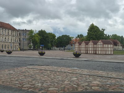 The image shows a view of a cobblestone plaza in Šilutė, Lithuania. In the foreground, there is a textured cobblestone path with some areas of darker, more uniform paving stones and a wider area of varied, irregularly shaped, reddish-brown and gray cobblestones. A dark gray, wet-looking paved road runs horizontally across the middle of the frame.
In the mid-ground, the plaza opens up, with several large, dark, circular planters filled with purple and yellow flowers. Behind the planters, there are various buildings. To the left is a multi-story building with a red tiled roof and a facade of many windows. Adjacent to this is a taller, gray building with a flat roof and windows. Further to the right, a distinct row of buildings with white walls and brown timber framing is visible, resembling traditional half-timbered architecture. Trees with lush green foliage are interspersed between the buildings, and some are visible in the background. A lamppost stands near the gray building.
In the distance, more buildings and trees are visible, suggesting a town or city environment. The sky above is overcast with heavy gray clouds, indicating a cloudy or possibly rainy day. There are no people or vehicles clearly visible in the scene. The overall impression is a historic or traditional urban plaza.