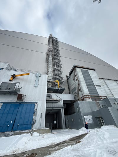 The image depicts an exterior view of the Chernobyl Nuclear Power Plant's New Safe Confinement (NSC) and adjacent structures, located within the Chernobyl Exclusion Zone, near the abandoned city of Pryp'yat', Ukraine. The scene is set on an overcast winter day, with a gray sky and snow covering the ground.

Dominating the background and upper portion of the frame is the massive, arched, silver-colored NSC, constructed from corrugated metal panels, covering Reactor 4. A tall, slender exhaust stack, featuring multiple external support structures, ladders, and yellow gantries or cranes, ascends vertically along the left side of the NSC.

To the left of the stack, a multi-level industrial building with light gray corrugated metal siding is visible. At its base on the left, large blue double doors, appearing to be loading bay entrances, are prominent. Above these doors, a yellow articulated crane arm is mounted to the building's facade, extending horizontally.

To the right of the central stack, an older, multi-story concrete structure with recessed dark window panels is partially visible, showing signs of wear. Closer to the foreground on the right, a lower, modern building with corrugated metal siding and a dark entrance is present.

The ground is uniformly covered in snow, with a cleared, dark gray paved pathway running diagonally from the bottom-center towards the mid-right, bordered by snowdrifts. One individual, wearing dark pants, a light-colored top, and a bright orange hard hat, stands on this cleared path in the mid-right, facing away from the camera towards the power plant structures. Overhead lights or floodlights are partially visible against the sky in the upper right corner.