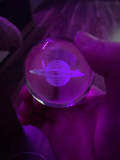 A close-up shot shows a hand holding a clear glass sphere, possibly a crystal ball. Inside the sphere, a three-dimensional representation of a planet, likely Saturn due to its visible rings, is etched or embedded. The scene is illuminated by a vibrant purple light, which casts a glow on the hand and the surrounding environment. The lighting creates a slightly ethereal or mystical atmosphere, with deep shadows and bright highlights. The hand's fingers are visible, gently cupping the sphere, suggesting care and perhaps wonder. The background is dark and out of focus, hinting at an indoor setting with little ambient light apart from the purple illumination. The overall impression is one of a captured moment of curiosity and appreciation for the miniature planet within the sphere. The location context is Gliwice, Poland, though no specific landmarks are visible to confirm this. There is no visible text in the image.