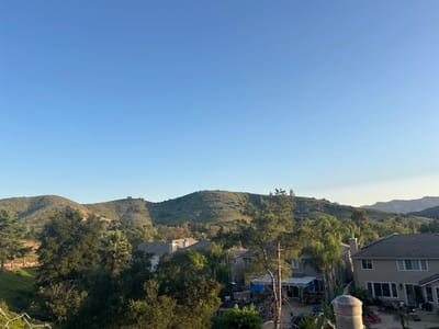 The image depicts a daytime view in Oak Park, United States of America, overlooking residential properties and rolling hills. The sky is clear blue. Several houses with tan exteriors and grey tiled roofs are visible among trees, obscuring the full view. Hills covered in vegetation form the backdrop. The immediate foreground shows foliage and a partial view of a fence.
