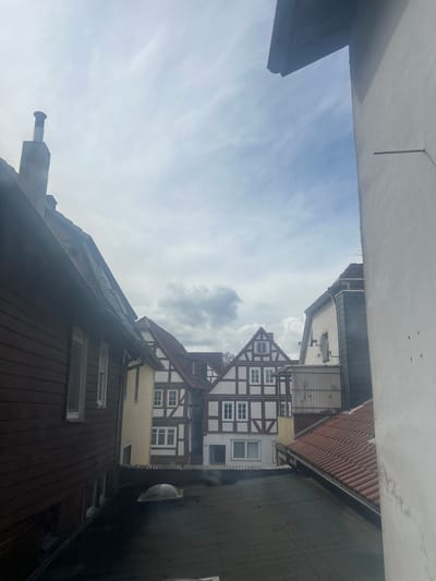 The image captures an elevated rooftop perspective within the city of Treysa, Germany, under a partially cloudy sky. In the left foreground, the dark brown, horizontal paneling of a building's facade is visible, equipped with white-framed windows and partially lowered external roller shutters. A grey concrete chimney with a silver-colored cylindrical cap projects from the dark roofline above this building. The immediate lower foreground features a dark, flat roof with a single translucent dome skylight.
On the right side of the frame, a plain white stucco wall extends vertically, displaying minor surface discoloration and a single horizontal metallic bar protruding from it. A reddish-brown tiled roof is partially visible in the lower right, descending from the white wall. The central midground and background are occupied by traditional German half-timbered houses, characterized by dark brown timber framing contrasting with white infill panels. These structures feature multiple white-framed windows and reddish-brown tiled gable roofs. One such building displays the address number "21" near its ground level entrance. Another building in the midground exhibits a section of grey slate tiling on its upper facade and a mounted satellite dish or antenna. A corrugated metal structure is situated between two of the central buildings. The sky overhead shows a mixture of white and grey clouds with visible patches of blue. No individuals are present in the scene.