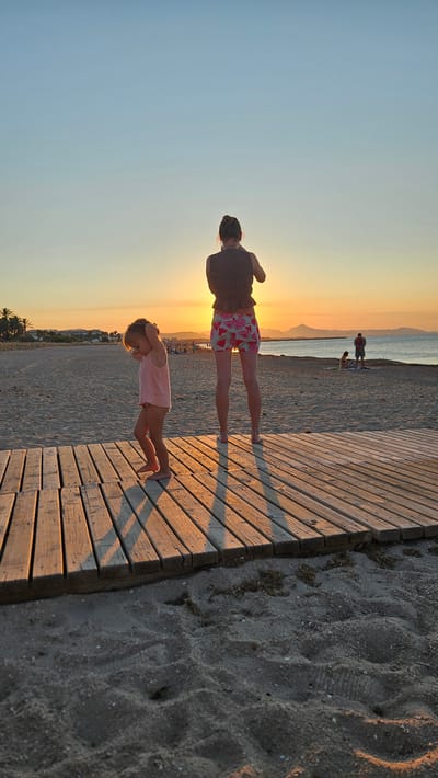 Two individuals, a woman and a young child, stand on a wooden boardwalk at the beach in Deveses, Spain, during sunset. The woman, viewed from behind, wears a dark, textured top and shorts with a watermelon pattern. She is holding up what appears to be a phone, possibly taking a picture. The child, also viewed from behind, is wearing a pink tank top and a diaper or similar undergarment. The child has their hands on their head and appears to be looking down. The boardwalk is constructed of wooden planks and extends across the sand. In the background, the sun is setting over the sea, casting a warm orange glow across the sky and the beach. Other beachgoers are visible in the distance, as well as palm trees and buildings along the coastline. The sand is textured with footprints and scattered debris.