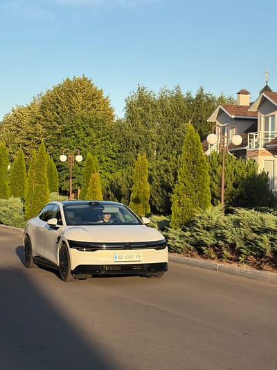 This is a vertical, slightly angled medium shot capturing a modern white car parked on a paved road. The car is the primary subject, positioned prominently in the foreground. Its sleek design and black accents are visible, with a license plate reading "AE 6587 YB" on the rear. A person is visible inside the car through the tinted windows, seated in the driver's position.

The setting appears to be a suburban or affluent residential area, with a manicured landscape of various evergreen trees and bushes lining the road. Behind the greenery, a multi-story building with a distinctive architectural style, featuring a brown roof and white trim, is partially visible. A double-headed lamppost stands amongst the trees, adding to the curated aesthetic.

The time of day seems to be late afternoon or early evening, indicated by the warm, long shadows cast by the car and the soft, golden light illuminating the scene. The sky is a clear, pale blue, suggesting fair weather. The overall mood is calm and serene. The location is identified as Slobozhanske, Ukraine.