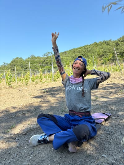 A woman with a braided ponytail and a blue baseball cap sits cross-legged on the ground in a vineyard. She is wearing a gray t-shirt with the words "HOW BRO" printed on it, blue overalls with black knee pads, and white athletic shoes. She has a pink neck gaiter around her neck and patterned sleeves on both arms, which appear to be tattoos or compression sleeves. Her right arm is raised with her fingers in a peace sign. Her left hand is on the back of her head. Behind her, rows of young grapevines are visible, supported by stakes and wires. The ground is covered with dry soil, stones, and sparse vegetation. A purple mat is visible to her right. The background shows a hillside covered with trees under a clear blue sky. The location is identified as Гурзуфский поселковый совет, Ukraine.