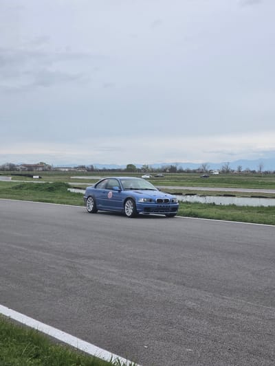 A blue passenger car is positioned on an asphalt race track next to a white painted track boundary line and a grassy verge. In the background is open agricultural land in the city of Kaloyanovo, Bulgaria under an overcast sky.