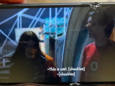 The image shows a close-up of a screen, likely a television or monitor, displaying a scene with two individuals and superimposed text. The screen has visible cracks and damage, particularly at the top and bottom edges. The background appears to be an illuminated, geometric structure, possibly indoors or an architectural element. In the foreground, two people are partially visible. The person on the left is mostly obscured by shadow, with long dark hair, and appears to be looking down. The person on the right is wearing a red shirt and is looking towards the person on the left. White subtitles are visible at the bottom of the screen, reading: "-This is cool. [chuckles]" and "-[chuckles]". The context of Wai A, Nigeria is not present in the visual information provided in the image.
