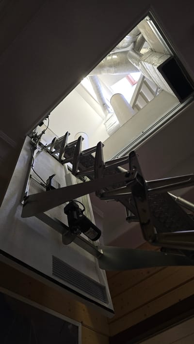 This is a low-angle shot looking upwards into an attic or crawl space. The primary subjects are a metal retractable attic ladder and ductwork. The ladder is extended, appearing to be made of aluminum and has a motor attached to one side, suggesting it is electrically operated. The ductwork, insulated with silver material, is visible above the ladder and extends into the space. There is also a white rectangular vent or grate visible on a white surface below the ductwork. The ceiling above is lined with wooden planks. Natural light is visible coming from a skylight or opening at the top of the frame, indicating it is daytime. The scene is indoors, likely a residential or commercial building, given the presence of ductwork and an attic ladder. No people are visible. No discernible text is present in the image.