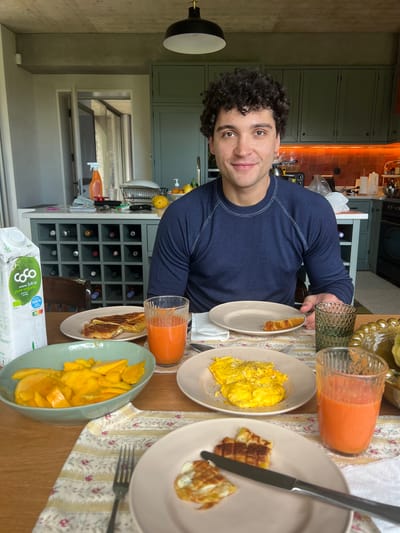A man with curly dark hair sits at a wooden dining table laid out with a breakfast meal. The table is covered with a floral print tablecloth. Dishes of scrambled eggs, waffles, mango slices, and slices of toast are arranged around the table. The man is wearing a long-sleeved navy blue shirt. He appears to be smiling at the camera. In the background, there is a kitchen area with light green cabinets, orange lighting, and a wine rack. A carton of coconut juice is visible at the edge of the table. The image may have been taken in Gondarém, Portugal, although there are no visual markers to confirm this location.