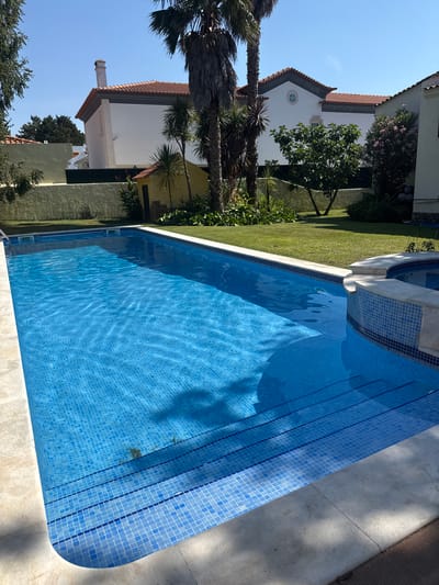 A tiled swimming pool dominates the foreground of this image, its bright blue water reflecting the clear, sunny sky. The pool has a gradual set of steps leading into it, also tiled in blue. The surrounding area is a lush green lawn, bordered by a light-colored patio.
In the background, a white house with a red-tiled roof is visible, featuring a prominent gable end with a small, round window. Palm trees and other greenery are scattered around the property, suggesting a warm climate. Another part of the house, or perhaps a separate structure, is also visible, with a more modern, flat roof. The scene is bathed in bright sunlight, indicating it is daytime. The overall impression is one of a serene and well-maintained residential property, likely in a coastal or Mediterranean-like location. There is no visible text in the image. The orientation of the image is such that the top of the frame is to the left.