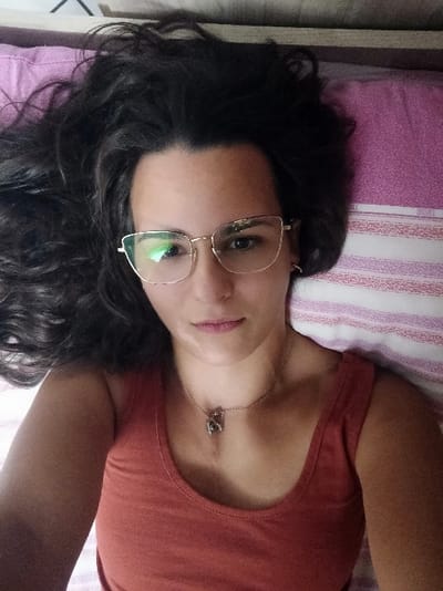 This image, located in San Marzano sul Sarno, Italy, depicts a female individual lying in a supine position. The subject, facing forward, has dark, wavy hair fanned out around her head against a pink and white horizontally striped pillowcase. She wears gold-framed, cat-eye style eyeglasses, with a green light reflection visible in the left lens. Her expression is neutral. A distinct area of lighter pigmentation is visible on her left jawline/chin area. She is wearing a rust-orange or terracotta-colored tank top. Around her neck is a thin silver-colored chain necklace with a small, dark, multi-faceted pendant. A dark, possibly wooden, edge is visible at the very top of the frame.