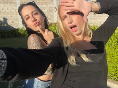 The scene is an outdoor residential or garden area located in Quilmes Oeste, Argentina. Two young adult females are depicted in a close-up, brightly lit photograph. The female on the left has dark hair, with sunglasses resting on her head, and a prominent mandala-style tattoo on her right upper arm. She wears a black top and light-wash denim jeans, with her left index finger touching her chin and lips pursed. The female on the right, positioned closer to the camera, has blonde hair, a septum nose piercing, and multiple bracelets on her right wrist. She wears a black cardigan over a black lace top. Her right hand is raised, fingers spread, shielding her face from direct sunlight, and her mouth is open. The background consists of a light-colored brick wall with an embedded outdoor light fixture, a manicured green lawn, and dense green shrubbery. Strong sunlight creates visible shadows on the grass and partially illuminates the subjects.