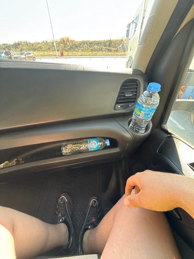This is a first-person view from inside a vehicle, likely a truck or van, showing the driver's perspective. The interior of the vehicle is visible in the foreground, including the dashboard with an air vent and two cupholders. One cupholder contains a partially filled bottle of water with a blue cap. Below this, in a recessed compartment, is another partially filled bottle of water. To the left of this compartment, on the dashboard, are several coins. The driver's legs and feet, clad in athletic shoes, are visible in the lower part of the frame, resting on the floor mat. A hand is resting on the driver's leg. Through the windshield, a bright, sunny outdoor scene is visible, showing a dirt road or parking area with several cars, a fence, and vegetation. The location is identified as Zagore, Bulgaria.