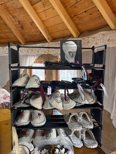 The image depicts an indoor market stall filled with handmade felt slippers and small pouches. The stall is a multi-tiered black metal rack. The top shelf holds a black and white photograph of a woman wearing a traditional headscarf, alongside some dark fabric pouches. The second shelf is primarily filled with colorful, embroidered small pouches with fabric loops. The third shelf features cream-colored felt slippers with embroidered designs, including a horse-like figure on one pair, and other slippers with elaborate trim. The fourth shelf displays more felt slippers, some with red embroidered roses, and more slippers with intricate scalloped trim. The bottom shelf shows a collection of felt slippers, some in a woven basket, and a plastic bag containing small stuffed animal figures, including pandas. The stall is set against a backdrop of a rustic wall, possibly made of adobe or earth, with white fabric curtains hanging in the windows. Above the stall, a wooden beamed ceiling is visible. The overall impression is of a display of traditional crafts in a local setting, likely a market or shop in the village of Кызыл-Туу, Kyrgyzstan.
