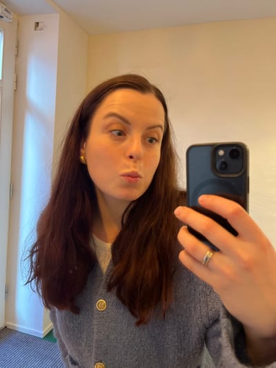 A female subject is depicted taking a mirror selfie indoors. She has long, dark brown hair and fair skin. Her attire includes a white undershirt beneath a textured, light grey cardigan fastened with large, circular gold buttons. She wears gold spherical stud earrings and a ring, appearing as two stacked bands, on the ring finger of her left hand. The subject holds a dark-colored smartphone, featuring a visible triple camera array, in her left hand, positioned to capture her reflection. Her lips are pursed in a pout or kiss, and her eyes are wide, directed towards the phone screen. The background consists of plain, light-colored walls, possibly off-white or light beige. A white door frame with visible metal hinges is present on the far left. A small, dark, rectangular recess or fixture is observed high on the left wall, above the door frame. Natural light illuminates the scene from the left. This scene is located in the city of Kaiserslautern, Germany.