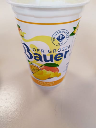The image shows a close-up of a white yogurt cup with a yellow and blue label. The label features a circular emblem with "PRIVAT-MOLKEREI" and "1887" printed on it, as well as an illustration of a person wearing a hat. Below the emblem, the text "DER GROSSE Bauer" is prominently displayed in large blue letters. The lower part of the label has an image of exotic fruits, including passion fruit and mango slices, and the text "EXOTISCHE FRUCHTE" in orange. The cup is placed on a light-colored, reflective surface, likely a table. The background is out of focus and appears to be a light, neutral color. There are no people or specific city landmarks visible that would allow for identification of a location in Bremerhaven, Germany.