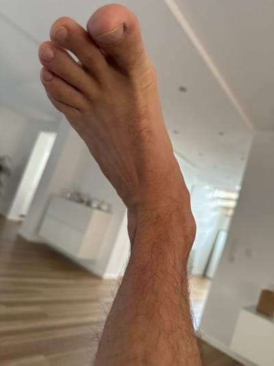 The image displays a close-up, low-angle shot of a person's bare left leg and foot, angled upwards and slightly to the right. The leg is covered in dark hair, becoming less dense towards the ankle and foot. The foot itself is pale and appears to be raised, with the toes pointed upwards. The background is blurred, suggesting an indoor setting. To the left, a wooden floor with horizontal planks is visible. In the mid-ground and background, elements of a modern interior are present, including white cabinets, a doorframe or wall, and what appear to be ceiling lights. The overall lighting is bright and diffused. The image does not contain any information that would allow for the determination of its location in Oberleichtersbach, Germany.