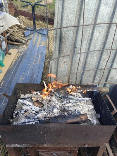 The image shows a metal brazier with burning wood inside. The brazier is mostly filled with ash and embers, but there are still some flames visible. The brazier is sitting outdoors, possibly in a backyard or garden. In the background, there is a metal fence and some other objects, including a table, chair frame and firewood. The location is Вичелово, Russia, based on the prompt.