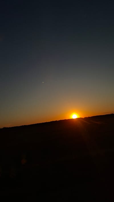 A landscape photograph shows a sunset. The sky on the left is dark blue, fading to yellow-orange in the middle where the sun is setting. The horizon line on the right is dark and indistinct, possibly trees or land. The sun is a bright orange orb. There's a small, pale dot in the sky on the left. The photograph appears to be taken at a tilted angle. The location is Любанскі сельскі Савет, Belarus.
