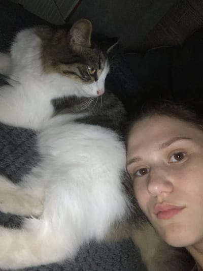In Belogorsk, Russia, a close-up, high-angle shot captures a young woman and a cat. The woman, on the right, has brown eyes and light skin, with her face angled towards the camera. Next to her, the cat, with white and tabby markings, is lying down, facing away from the woman and looking to the left. The background is dark and indistinct, suggesting an indoor setting. The composition emphasizes the shared space and proximity between the woman and her pet.