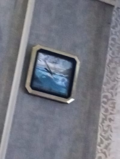 The image shows a framed picture of a blue ocean with a wave crashing. The picture is square with a light-colored frame. It is hanging on a textured gray wall. To the left of the picture is a vertical white trim. To the right of the picture, there is a section of wallpaper with a subtle floral pattern. The image is blurry and the quality is low. There are no people visible in the image.