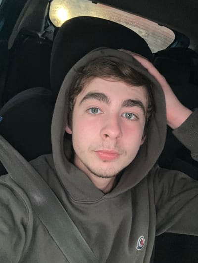 A young man with fair skin and light blue eyes is wearing a gray hoodie in a car. The hood is up, framing his face. He has dark brown hair and a light beard. A seatbelt is visible across his chest. The car's headrest is behind him. A car window with raindrops is in the background. The lighting suggests it might be dusk or evening. The man is looking directly at the camera with a relaxed expression.