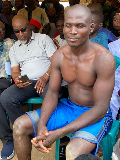 The image shows a group of people gathered in Kafanchan A, Nigeria. A shirtless man with a muscular build and blue athletic shorts is seated prominently. He is surrounded by other individuals, some of whom are also seated. One man wears sunglasses and a light-colored shirt, holding a phone. The overall environment appears to be outdoors, possibly a gathering of some sort. Many individuals are observed within this image.