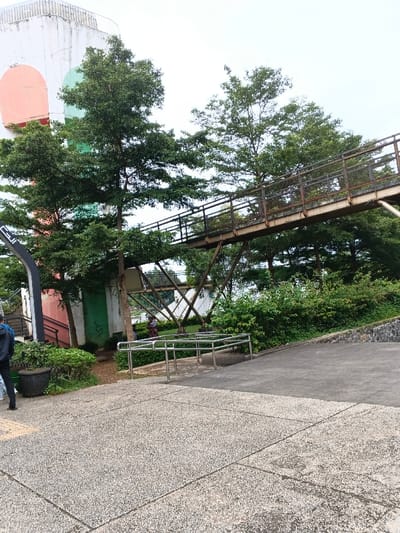 The image captures an outdoor scene, likely a park, campus, or recreational area, characterized by lush greenery, paved pathways, and a prominent elevated pedestrian bridge. The sky is bright and overcast, indicating daytime.
In the foreground, a light-colored, textured concrete path dominates the lower half of the frame. This path has visible expansion joints forming a grid pattern. On the far left, a person is partially visible from the back, wearing a dark backpack and dark clothing, with their right leg and foot visible as they appear to be walking away or standing. Next to this person, a large, dark-colored planter holds green, leafy shrubs. To the right of the person and planter, a set of silver-colored metal railings with multiple horizontal bars forms a section of a barrier or guide along the pathway, possibly leading to a ramp or steps not fully visible. Further to the right, the paved path changes texture and color, becoming a darker, smoother asphalt-like surface that slopes upwards towards the right edge of the frame, bordered by a low, rough stone retaining wall.
The middle ground is defined by a dense canopy of trees with vibrant green foliage, obscuring much of what lies beneath them. Interspersed among the trees is a weathered metal pedestrian bridge, stretching diagonally from the left side of the frame towards the upper right. The bridge appears to be constructed from rusty or dark brown metal, with a sturdy truss structure supporting its walkway. Its railings are also made of metal, featuring a pattern of vertical bars or mesh.
Behind the trees on the left, part of a tall, light-colored building or structure is visible. It features large, arched shapes painted in distinct colors – an orange arch and a green arch. A dark, possibly black, vertical structure with some indistinct white text (resembling "CUI" or similar characters) is also visible near the base of this building on the far left. Faint green graffiti marks are present on the light-colored wall section below the bridge on the left. Underneath the pedestrian bridge and further back, two small, indistinct figures, likely people, can be seen standing or sitting in a grassy area amidst more trees and shrubs.
The background primarily consists of the bright, overcast sky and the tops of more trees, extending into the distance. The very top of the white building on the left reveals what appears to be a cylindrical or circular structure with railings, possibly an observation deck or water tower. The overall impression is one of a well-maintained outdoor space with a mix of natural elements and human-made structures.