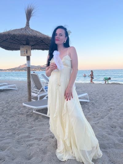 A woman with dark hair adorned with a white flower and wearing a pale yellow dress stands on a sandy beach. She is holding a delicate white accessory. Her red nail polish is visible on her hands. Behind her, the ocean stretches out with calm waves. In the background, several people are visible enjoying the beach and water. One man in a green shirt is standing near the water's edge, and another person in a bikini is walking along the shore. Further in the distance, mountains can be seen. The scene is illuminated by the soft glow of either sunrise or sunset, casting a warm light across the sky. Beach chairs and a straw umbrella are also present, suggesting a resort or public beach area. The overall atmosphere appears relaxed and serene.