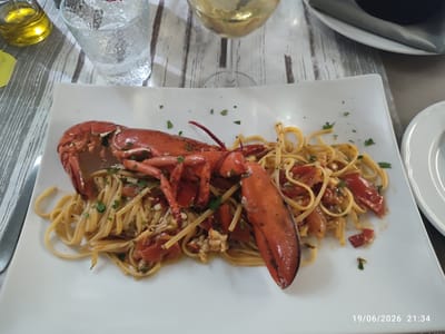 An overhead, eye-level shot shows a white rectangular plate with a lobster pasta dish and a glass of white wine on a wooden table with a striped placemat. A whole cooked lobster sits atop a bed of linguine with cherry tomatoes and chopped parsley. The lobster is bright red, with claws and tail clearly visible. The pasta is light yellow and appears to be tossed in a light sauce. The wine is a pale yellow color and is in a stemmed glass. The table surface is made of light-colored wood with a distressed finish. The placemat is gray and white striped. A bottle of olive oil is visible in the upper left corner of the image. The bottom right corner of the image contains a timestamp: "19/06/2026 21:34". The scene appears to be a restaurant setting in Scordia, Italy.