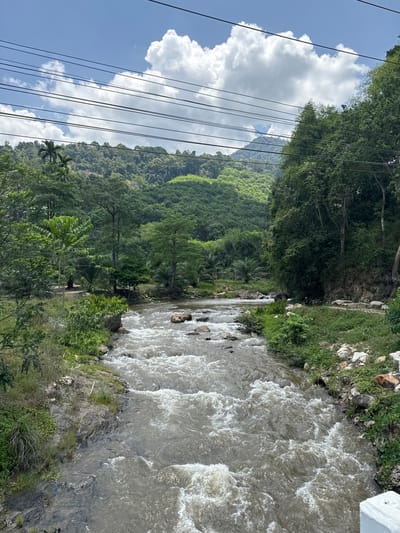 The image shows a river flowing through a lush, green landscape in Tambon Song Phraek, Thailand. The river is turbulent, with white water visible amongst the grey current. Vegetation lines both banks of the river, including trees and smaller plants. A mountain covered in dense foliage is visible in the background, framed by a partly cloudy sky with power lines.