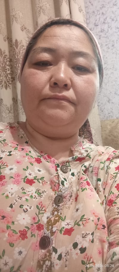 A woman with fair skin and graying dark hair tied back in a pink headscarf poses for a selfie. Her eyes are dark and slightly closed, and her lips are pursed. She wears a beige blouse with a floral pattern of pink, red, and white flowers. Two dark, round buttons are visible on the front of her blouse. The background consists of a patterned curtain with a beige and gold damask design on the left and a textured wallpaper on the right. The lighting suggests an indoor setting. The image is dated 24.06.2026 08:24.