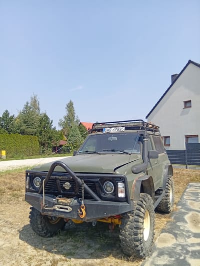 A modified, olive green, four-door Nissan Patrol SUV is parked outdoors on a sunny day. The vehicle features large, aggressive off-road tires, a winch mounted on the front bumper, a snorkel, and a roof rack with a light bar. The SUV is positioned on a dirt and gravel surface next to a paved area with a textured pattern. In the background, a modern, light-colored house with a dark roof and a black metal fence is visible. Dense evergreen trees and a trimmed hedge line the property, with a small yellow utility box located near the hedge. The license plate on the vehicle reads "DWR 69885". The image appears to have been taken in Długołęka, Poland, based on the license plate format. There are no people present in the image.