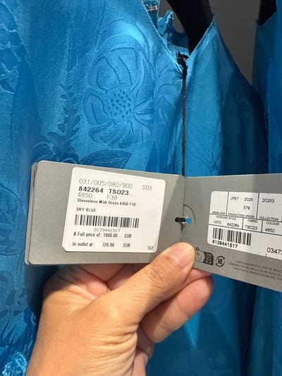 A person's hand is holding up a price tag in front of a bright blue, textured dress. The tag displays the dress's product code (842264 TS023), size (F38), and description ("Sleeveless Midi Dress 4850 F38 SKY BLUE"). It also shows a full price of 1800.00 EUR and an outlet price of 720.00 EUR, along with a barcode and other product information. The image is taken indoors, likely in a retail environment, and no specific location in the city of La Roca del Vallès, Spain is visible or identifiable.