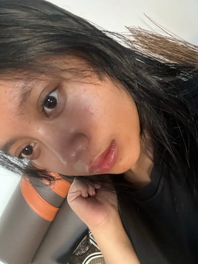 A close-up, angled, selfie-style photograph shows a young woman's face, primarily focusing on her eyes, nose, and mouth. Her dark hair falls across her face, partially obscuring her forehead and left eye. She appears to be wearing a black t-shirt. The background is out of focus but suggests an indoor setting with a white wall on the left and a brown and orange textured surface, possibly upholstery, on the left side of the frame. The overall lighting is soft, highlighting the sheen on her skin and the gloss on her lips. The image does not provide any information about the specific location within Jambearum, Indonesia.