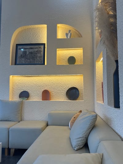 The image depicts a well-lit indoor scene, possibly a living room or lounge area, with a focus on textured white walls featuring recessed niches and shelving. The overall aesthetic is modern and minimalist, with warm, ambient lighting emanating from the niches.
On the wall, there are several decorative elements. One niche contains a framed black and white artwork. Another niche holds a round, dark decorative plate. Above this, a curved, terracotta-colored object is placed. Further to the left, a smaller niche displays a light green decorative plate with a leaf pattern. In another arched niche, a white sculptural object resembling a bird or a dove is visible. At the very top, a dark, elongated object, possibly a decorative piece or part of the ceiling structure, is present. A fluffy, dried plant decoration hangs from the ceiling.
To the right side of the frame, a large, light-colored sectional sofa is partially visible. A light grey throw pillow rests on the sofa, and peeking out from underneath it is a plush toy with a round face and small brown ears, giving a touch of whimsy to the otherwise sophisticated setting. The sofa appears to be made of a smooth, possibly leather or faux leather material.
The lighting suggests it could be evening or a dimly lit day. The scene conveys a sense of calm and contemporary interior design. There is no visible text in the image. The location context of Việt Trì, Vietnam, suggests this might be a home or a commercial establishment in that region.