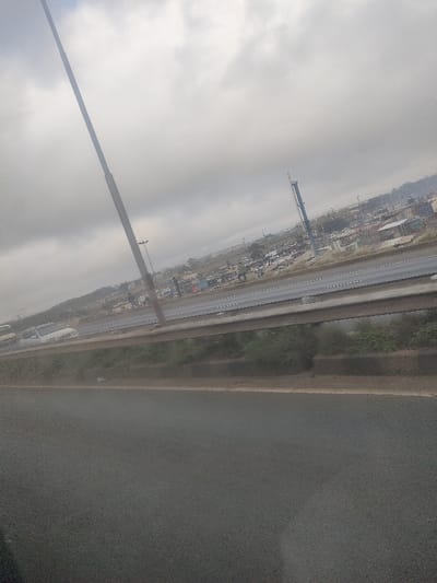 The image depicts a roadside scene in Biashara ward, Kenya, under an overcast sky. In the foreground, a dark asphalt road surface occupies the lower third. Immediately above this, a metal crash barrier runs horizontally across the frame, with green vegetation visible directly behind it. Beyond the barrier, a multi-lane highway stretches horizontally, featuring white lane markings. Several vehicles are present on this highway, including a white vehicle partially visible on the left. Multiple individuals are observed walking along the shoulder or an adjacent pathway of this highway. A tall, dark utility pole stands prominently on the left side, extending from the bottom of the frame towards the top. In the background, beyond the highway, a dense settlement of numerous structures, possibly informal housing with various roof types including corrugated iron, is visible. The landscape ascends towards distant hills. A tall, slender tower, possibly a communication mast, is present on the right side of the background within the settlement. The sky is heavily clouded.
