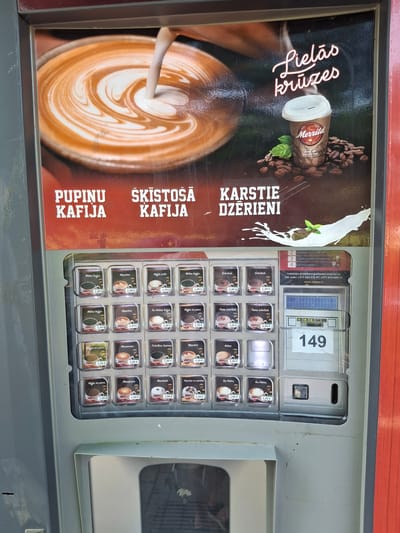 The image shows a Merill coffee vending machine located in Lēdmane, Latvia. The machine is in a public area, likely outdoors, with a red-colored wall visible on the right side. The top portion of the machine features an advertisement with an image of latte art and a cup of coffee. The advertisement includes text in Latvian: "Lielās krūzes" (large cups), "Pupinu kafija" (bean coffee), "Šķīstošā kafija" (instant coffee), and "Karstie dzērieni" (hot drinks). Below the advertisement is a display of various coffee and hot drink options with their prices, indicated in Euros. To the right of the drink selections, there is a control panel with a digital display showing "149", a coin slot, and buttons. The machine appears to be in working order.