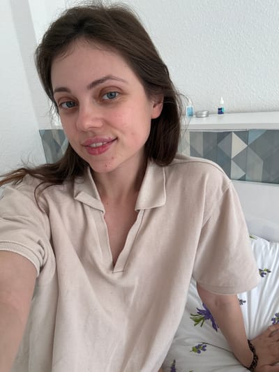 A young woman with fair skin, brown hair, and blue eyes is smiling at the camera. She is wearing a beige collared shirt. She is lying on a bed with a white floral-patterned pillow. Behind her is a white wall with a geometric patterned panel. The specific location of Kőröshegy, Hungary is not discernible from the image.