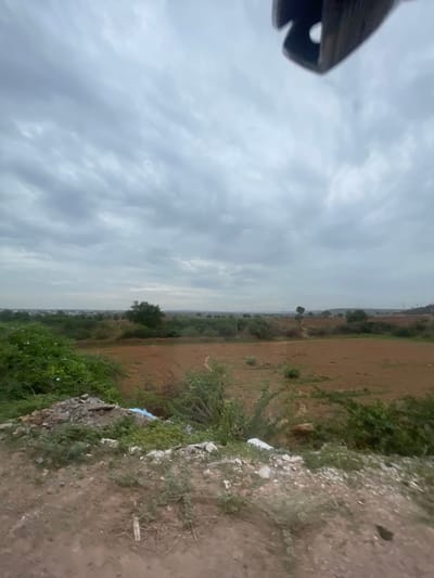 A wide-angle shot captures an overcast sky over a rural landscape in Bethamcherla, India. In the foreground, a pile of debris, including rocks and possibly plastic, sits on a dirt embankment covered with sparse vegetation. Beyond the embankment, cultivated fields in various shades of brown and green stretch towards a distant horizon dotted with scattered trees and faint structures. The overall impression is one of a utilitarian or undeveloped area, likely on the outskirts of a town or village.