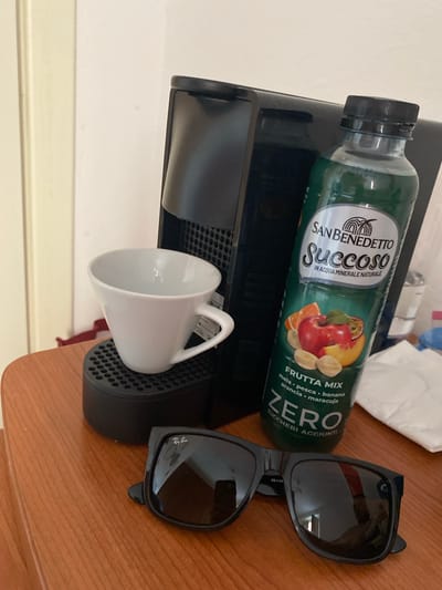 The image shows a black Nespresso coffee machine with a white espresso cup on its drip tray. To the right of the coffee machine is a bottle of San Benedetto "Succo" Frutta Mix juice. In the foreground, a pair of black Ray-Ban sunglasses are placed on a wooden surface. The background is a plain white wall. The scene appears to be set indoors, possibly in a hotel room or a domestic setting, on a wooden table. The image does not provide any visual clues that would suggest a specific location within the city of Cagliari, Italy.