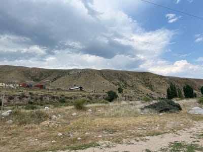 The image depicts a rural landscape with rolling hills in the background. In the middle ground, there are a few structures, including a house or small building with a dark roof, some shipping containers, and a cluster of tall, slender trees. The foreground is a dry, grassy field with scattered rocks and some sparse vegetation. A pile of pruned branches is visible on the right side of the foreground. The sky is partly cloudy with patches of blue sky and heavier gray clouds. The overall impression is one of a remote, undeveloped area. This scene is located in the village of Yuryevka, Kyrgyzstan.