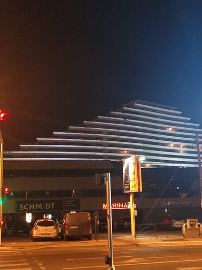 This image captures a night scene in front of a terraced building, likely a commercial or residential complex, in Villeneuve-Loubet, France. The building's architecture features multiple stacked levels, creating a stepped or tiered effect, with white horizontal bands delineating each level. Some windows on the upper floors are illuminated, suggesting occupancy.
At street level, the building houses several businesses. A prominent sign reads "SCHM DT", likely part of a store name. Adjacent to it, another illuminated sign displays "MARINA". Further to the right, a sign indicates "maxi ZOO".
Several cars are parked in front of the businesses, including a white hatchback, a dark van, and other vehicles partially visible. A traffic light with a red signal is visible on the left side of the frame, and a pedestrian crossing is marked on the street. A street lamp casts a warm glow on the scene, and the overall ambiance is that of a well-lit urban environment at night. A signpost with multiple advertisements is also present in the foreground. The sky is dark, indicating nighttime.