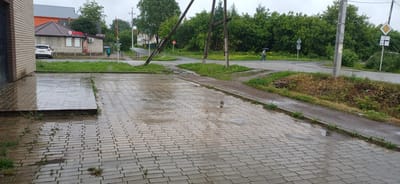 This is a ground-level, wide shot of a wet, paved area in front of a building, likely after rain. The paving stones are a light grey color and reflect the overcast sky. In the background, a road intersection is visible with a crosswalk. A person holding a blue umbrella is walking across the intersection, and a white car is parked to the left of a small building. The building has a brown roof and brick facade with white window coverings. To the right of the intersection, there are trees and various road signs, including a yellow diamond-shaped sign with a black border. The scene appears to be in Zuevka, Russia, given the context.