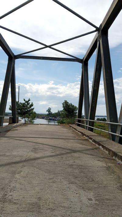 The image shows a view from a concrete bridge under a cloudy sky in Baroh Lancok, Indonesia. The bridge has a dark metal truss structure overhead. The road surface is cracked concrete. On either side of the road, there are concrete barriers with metal railings. In the distance, a body of water is visible with several boats docked. There are trees and buildings on the far bank of the water. The scene appears to be a rural or semi-rural area. There are no people visible in the image.